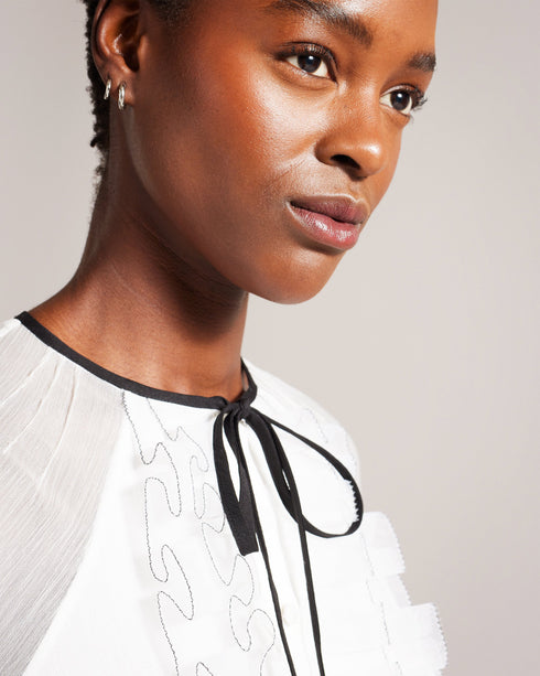KELEIH-Tops-Channel vintage glamour with a modern twist in the KELEIH blouse. Its delicate organza ruffles and darts exude timeless elegance, while the crew neck keeps it versatile. Whether you're attending a formal event or dressing up for a date night, this blouse will effortlessly elevate your look. Blouse Organza ruffles on front Long sleeves Contrast stitching and neck tie Button fastening Darts on shoulders Crew neck with contrast binding Silvia is 5'11" / 1.8m and wears White in a Ted size 1- Ted Bak